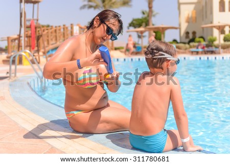 Mother and her young son sitting on the surround of a resort swimming pool putting on sunscreen to protect their skin while enjoying the summer sun