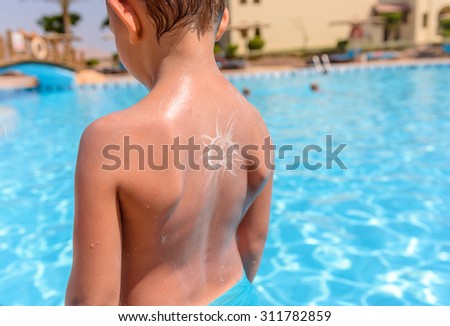 Young boy with a fun artistic sunscreen sun drawn on his back by his mother standing overlooking sparkling sunlit water in a swimming pool