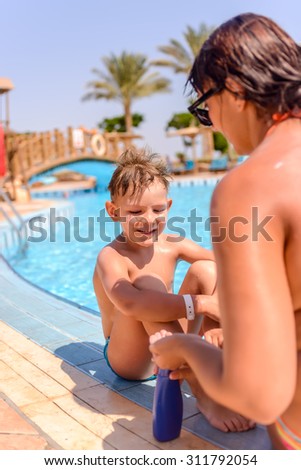 Woman applying sunscreen to the face of her young son as he sits waiting to go swimming alongside a swimming pool, close up of him looking at the camera