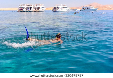 Man skin diving in the ocean lying on his back waving at the camera with diving tour boats in the background
