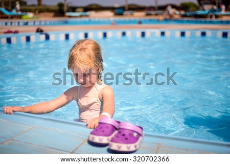 Cute little girl paddling in the kiddies pool at a resort standing in the water holding the edge of the tiles with her pink shoes in the foreground