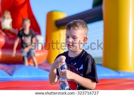 Young boy drinking bottled water as he stands in front of a colorful plastic jumping castle at a playground or fairground