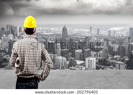 rear view technician builder in protective safety equipment goggles hard hat and and blueprints against balcony overlooking city dusky before rain falling