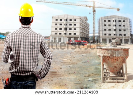 rear view of technician builder in protective safety equipment  hard hat and and blueprints at the front of blurred construction site