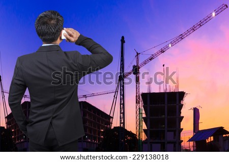 rear view business man in suit speaking mobile phone looking to the future against silhouette of crane and building construction and beautiful sunset sky