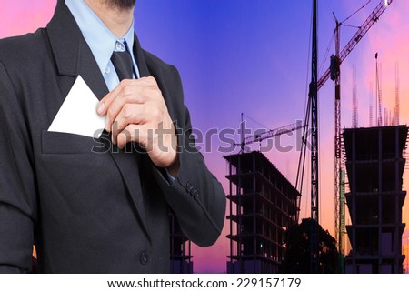 business man takes out business card from the pocket of business suit against silhouette of crane and building construction and beautiful sunset sky