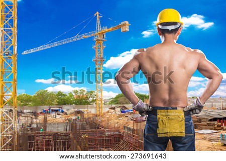 a back view of construction worker showing his muscles at footing of building construction site with tower crane and blue sky
