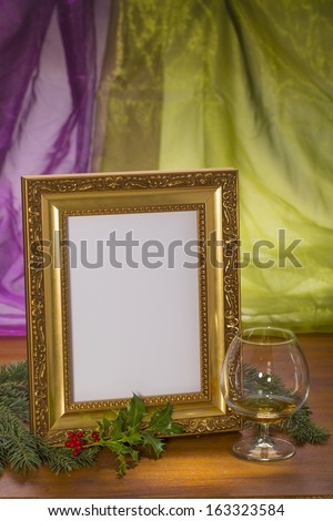 golden frame with brandy glass and evergreens on wooden in front of purple and green background