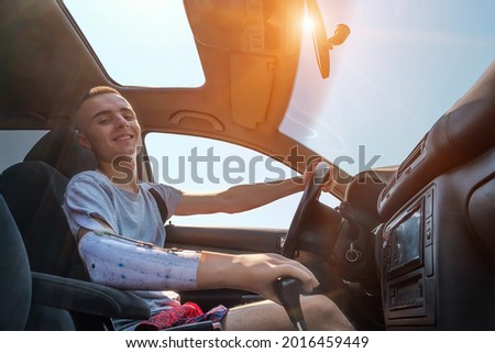 Young smiling caucasian man with amputated arm and prosthesis drives a car on a sunny day in summer. Side view inside of cabin Imagine de stoc © 
