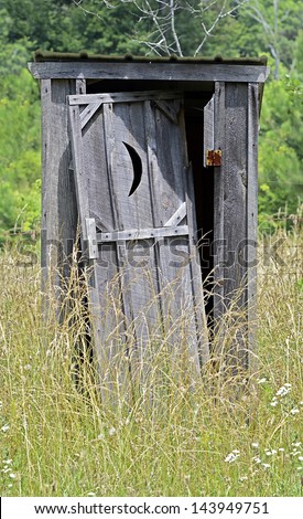 An old outhouse with door falling off surrounded by weeds.