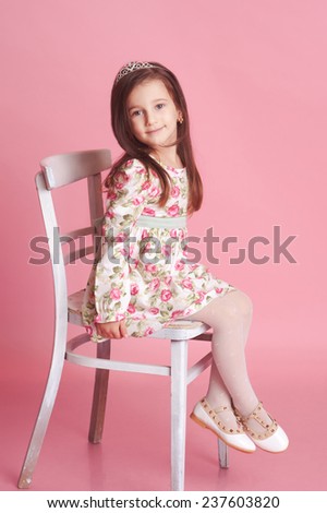 Smiling baby girl 4-5 year old posing in studio over pink. Sitting on chair. Wearing trendy floral dress.