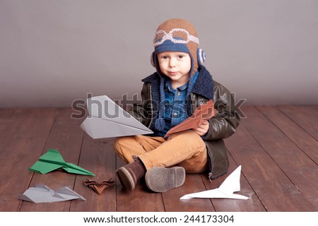 Cute baby boy wearing stylish leather jacket and aviator cap over gray. Playing with paper origami planes in room. Sitting on wooden floor. Looking at camera. Childhood.