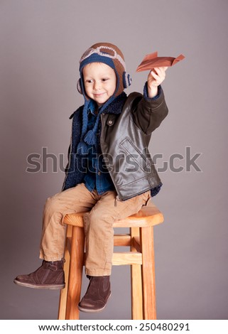 Cute baby boy holding with origami plane in room over gray. Wearing stylish leather jacket. Smiling kid. Childhood. Sitting on wooden chair.
