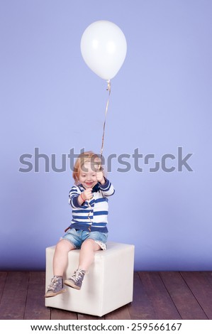 Child girl 2-3 year old holding white air balloon in room. Sitting on chair over purple. Wearing striped shirt and denim shorts. Playful baby. Childhood.