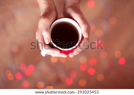 Woman holding mug with black hot tea at glowing light background. Colorful. Christmas decorations. Top view.