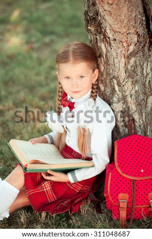 Smiling school girl 6-8 year old reading book outdoors. Wearing stylish school unifom. Looking at camera. Childhood. Back to school.
