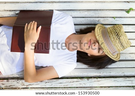 Young woman lies on a bench with a book covered with a hat, top view