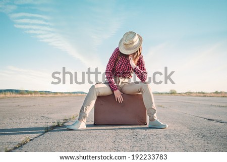 Hipster woman in the hat sits on suitcase on road
