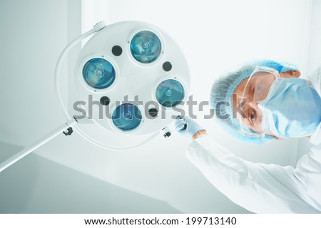 Man surgeon in protective uniform places a surgical lamp for operation and looks at camera. Focus on surgical lamp