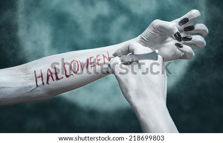 Unrecognizable dead woman writes the word Halloween by scalpel on her arm on background of full moon. View of human arms. Halloween or horror theme