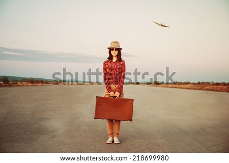 Traveler woman stands on road with vintage suitcase, airplane in sky. Space for text