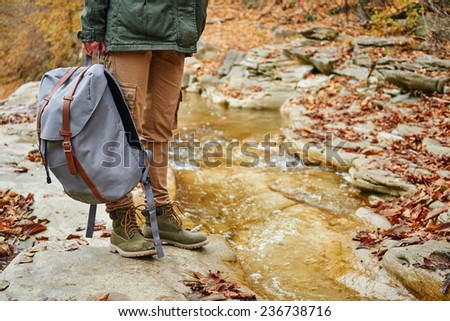 Unrecognizable female hiker with backpack standing near a stream in autumn forest, view of legs. Focus on backpack