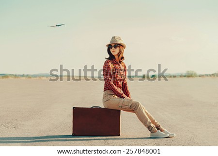 Traveler young woman in a hat sitting on suitcase on road. Airplane flying in the sky