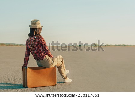Traveler young woman in a hat sitting on suitcase on road. Space for text in left part of image
