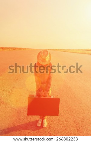 Traveler woman in hat stands on road with vintage suitcase, face is not visible. Space for text in the upper part of image. Image with sunlight effect