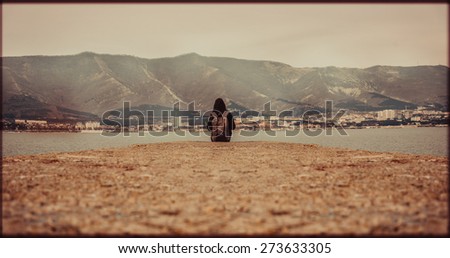 Traveler woman with backpack sitting on pier at bay in front of mountains. Space for text in lower part of image. Image with vintage color effect and rectangular frame