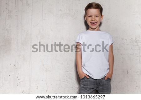 Cute little boy in white T-shirt posing in front of grunge concrete wall. Portrait of fashionable male child. Smiling boy posing, gray wall on background. Concept of children style and fashion. ストックフォト © 