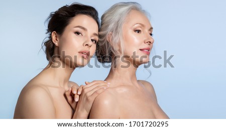 Elderly woman and young woman with perfect skin portrait. Young daughter standing behind older mother putting hand on arm looking at camera. Different age generation family bonding. Beautiful people