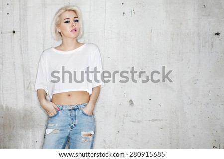 Naughty fashion young blond woman in a white T-shirt and jeans posing against the backdrop of a cement wall