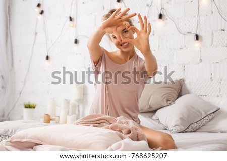 Photo of Beautiful young woman sitting on bed and shows gesture heart with fingers. Portrait of smiling lady posing in bright home apartments. Concept happiness and love.