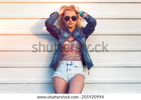 Outdoor summer closeup portrait of young stylish fashion glamorous woman or girl posed in sunny day on street jeans jacket and sunglasses standing near white wall