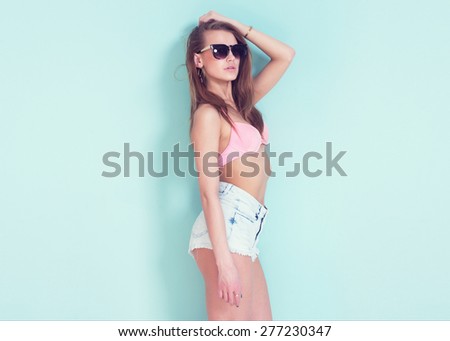 Outdoor summer closeup portrait of young stylish fashion glamorous sexy brunette woman or girl posed in sunny day jeans shorts swimsuit bra and sunglasses standing near blue wall.
