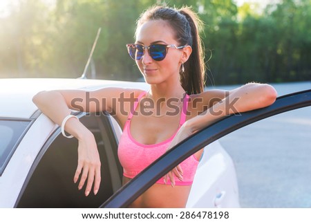Athlete sporty fit young woman in sports bra wearing sunglasses standing leaning on car with door open looking at camera