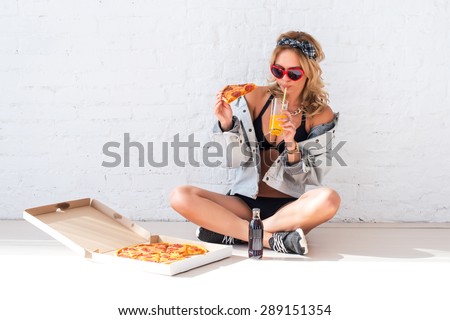 Young woman eating a piece of pizza drinking juice sitting on floor wearing sunglasses.