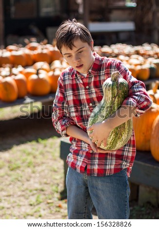 vertical orientation of boy with autism and down\'s syndrome in a bright red plaid shirt holding a large squash in his arms with pumpkins in the background / Visit to the Farm