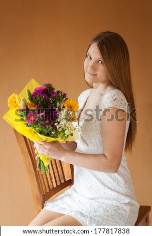 vertical orientation of a lovely, smiling, teenage girl in a white dress holding a colorful bouquet of flowers as she sits on a chair, with a warm, neutral background / Flowers say Congratulations