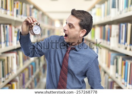 Man wearing a blue shirt and red tie. He is looking surprised to a clock. Over library background