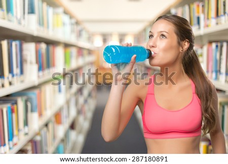 Young woman wearing gym clothes. She is drinking from a blue bottle. Over library background