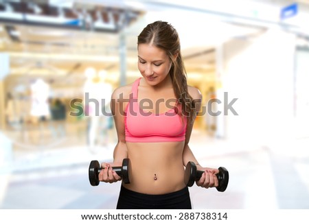 Young woman wearing gym clothes. She is holding dumbbells. Over shopping center background