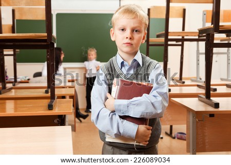 Schoolboy is in the half empty classroom after classes. He holds copybooks in his hands. He is bored. Teacher is teaching schoolgirl behind him.