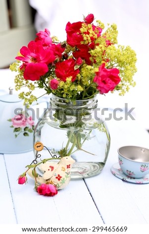 Re-cycled glass jar with wild red roses and green Lady\'s Mantle flowers , with shabby chic tin tea pot and cup and bird ornament on pale blue wooden floorboards, summer outdoor wedding  pretty picnic