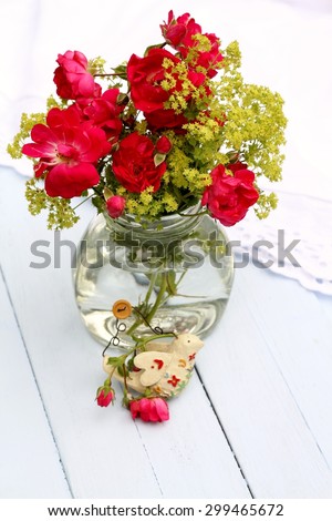 Re-cycled glass jar with wild red roses and green Lady\'s Mantle flowers , with shabby chic   bird ornament on pale blue wooden floorboards, summer outdoor wedding  pretty arrangement,