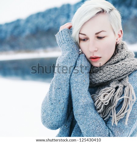 Outdoor winter fashion portrait of young attractive woman in park near the river. Cold blue colors. Girl with red lips dressed in sweater and scarf.