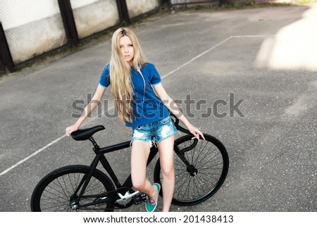 Pretty blonde serious fashion girl in jeans shorts posing in urban grey style square with her black fixed gear sport bicycle