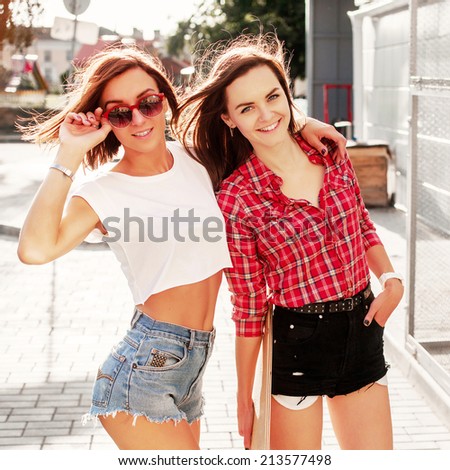 Outdoor colorful vintage portrait of two pretty fashion girls friends dressed in sexy shorts sensual photo with longboard urban style smiling and laughing in evening back light