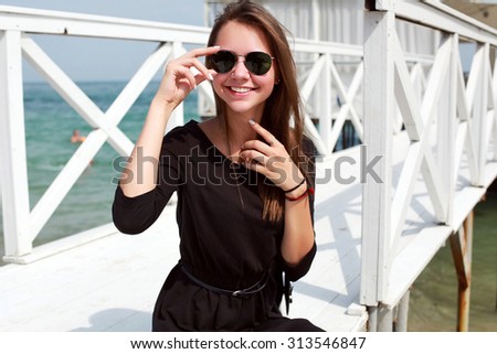 Pretty young brunette happy woman posing outdoor in summer feels happy sitting on white wooden pier in black dress